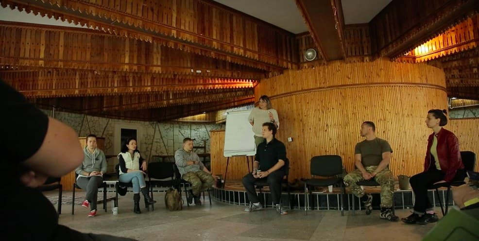 A training group sits in a wide circle in a wood-panelled hall.