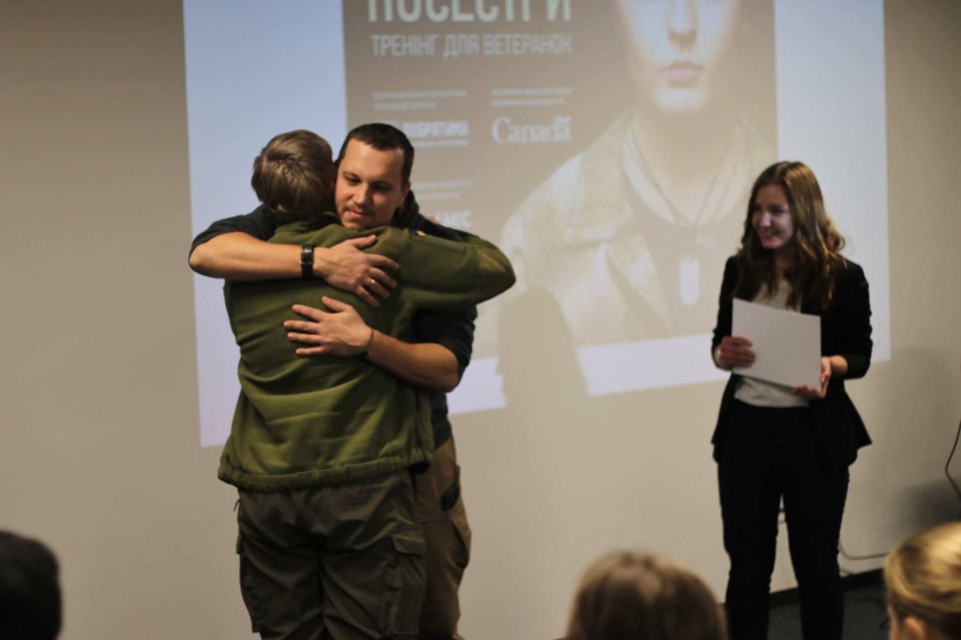 A veteran in uniform embraces a trainer at the close of a training session, while a colleague holding a certificate smiles.