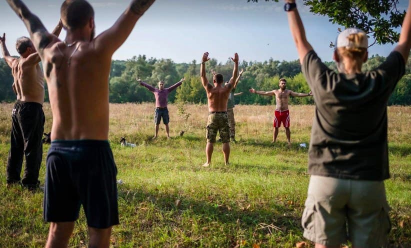 A group of veterans stands in a meadow with their arms raised above their heads.